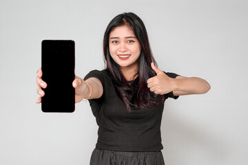An Indonesian woman in a black outfit smiles while holding a smartphone towards the camera and giving a thumbs-up. She appears happy, promoting or recommending something.