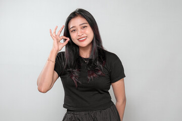 Fototapeta premium A cheerful Indonesian woman in a black t-shirt makes an OK hand gesture while smiling. She radiates positivity, confidence, and happiness in a modern studio portrait.