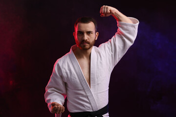 Man wearing uniform practicing karate in color lights and smoke against black background