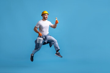 Man wearing hardhat with brush and bucket of paint jumping on blue background