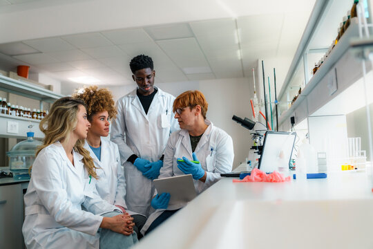 Professor holding a tablet, explaining scientific research concepts to a diverse group of students in lab coats, collaborating in a university laboratory focused on learning and innovation