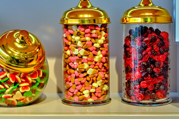 Various sweets: candies, lollipops, gummy worms, in a glass jar with a golden lid on the counter in...
