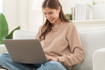 Fototapeta premium Concentrated Asian girl sit on couch working on laptop browsing internet at home, young woman busy using computer surfing wireless web shopping online
