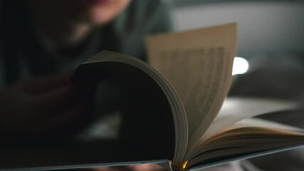 A man flips through the pages of a book in a dark room on his bed.
A man reads a book in the evening light.
A man relaxes reading an interesting book in a soft and cozy bed before going to sleep