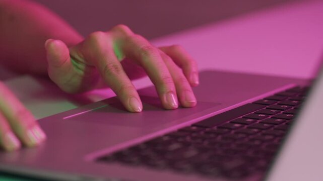 Close up shot of hands of unrecognizable person interacting with laptop touchpad in dimly lit environment with pink lighting