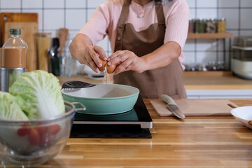 An old woman is dropping eggs into a pan, she is in the kitchen.