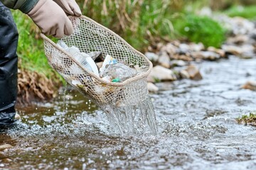 A person bends over the stream, holding a net full of collected trash. Water drips from the gloves as they participate in an environmental clean-up initiative