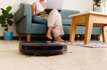 A black robot vacuum cleaner cleaning the floor and a woman with a laptop sitting on a sofa at home.