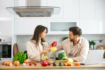 A couple joyfully prepares a colorful meal in a bright, modern kitchen filled with fresh vegetables and ingredients.