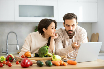 A couple leans over a laptop at the kitchen table, showcasing their collaboration amidst fresh vegetables and kitchen elements.