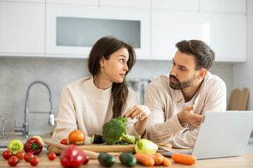 A thoughtful couple in a bright kitchen reflects on cooking options while engaging with fresh vegetables like broccoli and carrots.