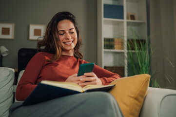 Smiling woman using smartphone and reading book at home