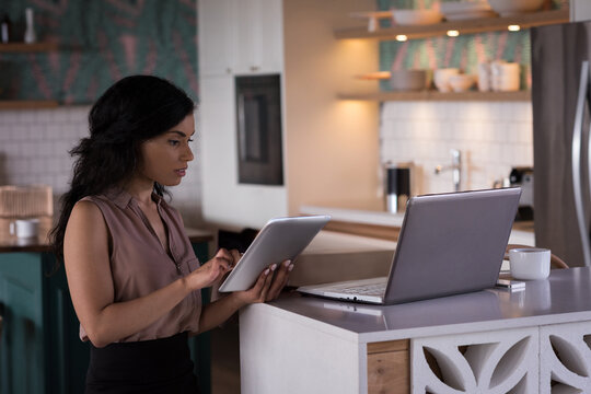 Using tablet and laptop, professional woman multitasking efficiently in modern kitchen, copy space