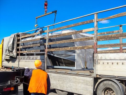 Workers are carefully loading large condenser units onto a flatbed truck on a sunny day.