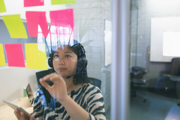 Asian woman using headset and tablet, organizing colorful sticky notes in office, copy space