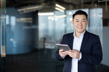 Portrait of a smiling young Asian man in a suit standing in the office lobby, holding a tablet and looking at the camera