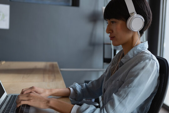Asian woman wearing headphones typing on laptop at office, focused on work, copy space