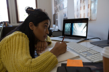 Asian woman studying at home, writing notes while watching laptop screen attentively, copy space