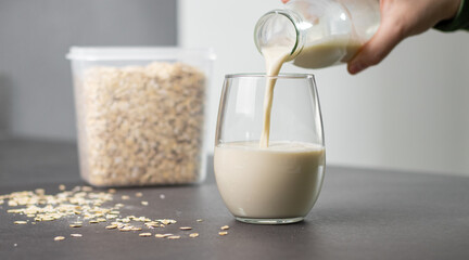 Close up of a female hand pouring oat milk into a glass on grey kitchen table. Vegan and non-dairy alternative milk