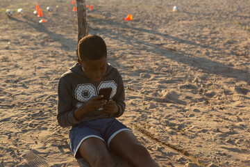 Young boy sitting on sandy field using smartphone, surrounded by soccer cones, copy space