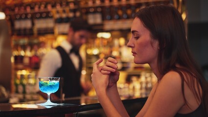 Sitting beautiful woman guest feeling bored waiting friends or couple with drinking mixed vodka in blue hawaii cocktail at luxurious counter bar for Friday meeting on bartender background. Vinosity.