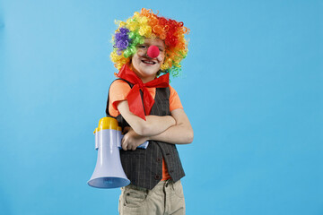 Happy little boy dressed like clown with megaphone on light blue background. Surprise party