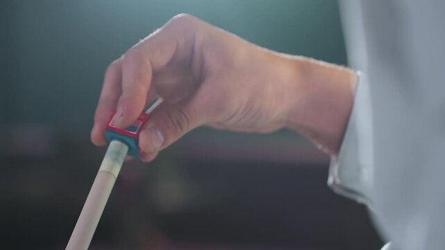 Close-up shot of hand in white shirt applying red and blue chalk to cue stick before billiards game. Focused grip and blurred background emphasize preparation for the next move