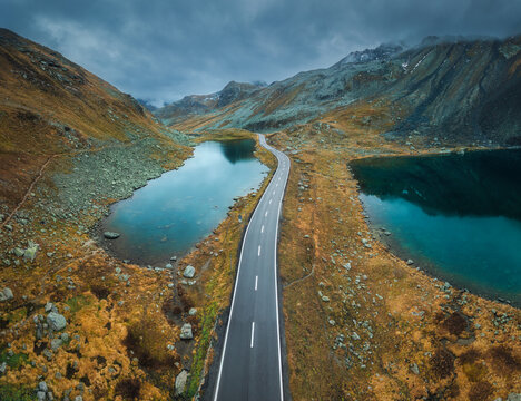 Aerial view of mountain road between two lakes in alpine valley in cloudy autumn day in Switzerland. Top drone view of empty road in swiss mountain pass, orange grass, ponds, overcast sky in fall