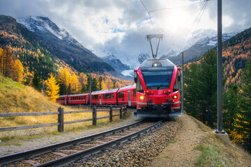 Red modern electric train traveling along a railway, winding through a stunning high mountains with snow-capped peaks and vibrant autumn trees in switzerland. Bernina Express in fall. Railway tourism © den-belitsky