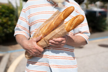 Old man with a long white beard holding two fresh baguettes wrapped in paper while wearing a striped shirt outdoors on a sunny day
