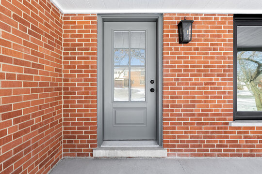 Fototapeta A home's grey front door with frosted glass surrounded by red brick, white ceiling, concrete patio, and black framed window.