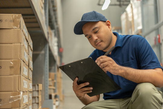 Warehouse worker wearing a hat and black shirt checking quality by clipboard on tall shelves in warehouse storage. Asian auditor or staff work looking up stock taking inventory in warehouse store.