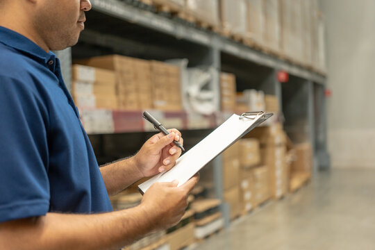 Warehouse worker wearing a hat and black shirt checking quality by clipboard on tall shelves in warehouse storage. Asian auditor or staff work looking up stock taking inventory in warehouse store. - Powered by Adobe