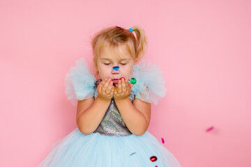 Girl with long hair in festive paper hat and dressy dress on pink background tossing gold confetti