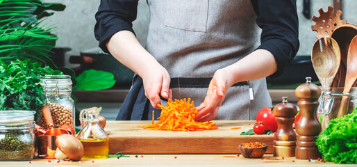 Cook blogger holds sliced ​​carrot on the blade of knife.. Cozy kitchen with wooden table, kitchenware, vegetables, herbs and ingredients for cooking. Healthy vegan food, culinary, recipes