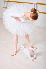 Young female ballerina bends gracefully towards crawling baby girl on wooden dance floor. Both wear...