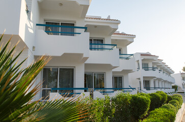 Obraz premium White resort exterior under bright sunlight features large balconies and lush green hedges. Modern architectural design with clean lines viewed at angle enhances its appeal