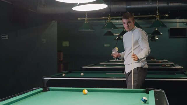 Man in white shirt and grey trousers holds cue stick and glass of lemon drink while pacing around billiards room. Overhead lights cast a moody ambiance, enhancing the stylish and relaxed atmosphere