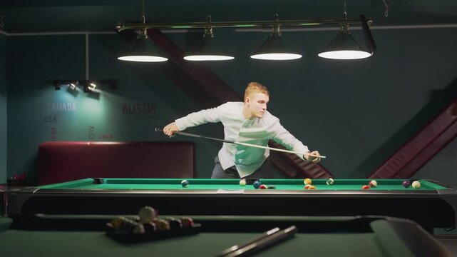 Young man in white shirt and grey trousers leans over green pool table, gripping cue stick with intense focus as he lines up shot. Overhead lights cast glow in stylish billiard hall with red seating