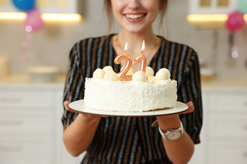 Coming of age party - 21st birthday. Happy young woman holding tasty cake with number shaped candles at home, closeup