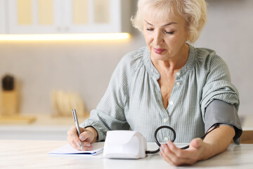 Senior woman writing results of blood pressure measurement in kitchen at home. Space for text