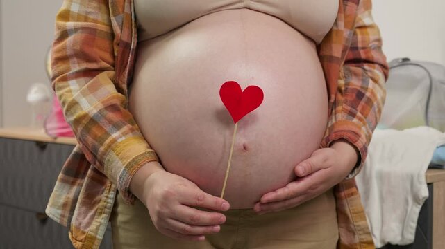 Pregnant woman holding red heart shape and feel love for unborn baby in belly. Awaiting for baby. Concept of maternity and pregnancy care