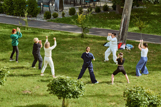 Adults, diverse ethnicities, practice qigong on grass under sunny skies. Instructed by mature woman in white, serene setting enhances focus and harmony