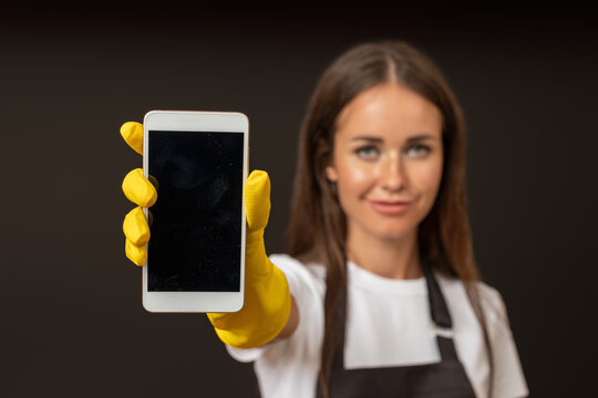 A female cleaner in a black apron and yellow rubber gloves, holding out a smartphone towards the camera, standing against a dark background.