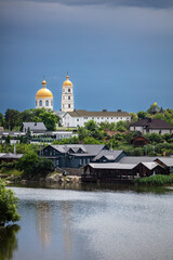 A picturesque town with a river in the foreground, lush greenery, and golden-domed churches in the background. The dramatic sky adds contrast, creating a serene yet moody atmosphere.