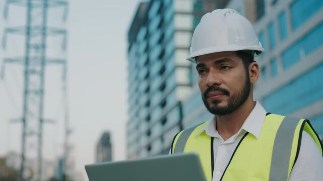 Portrait of skilled mixed race contractor working on laptop against modern office buildings. Mature bearded engineer uses software to control construction site. Building technology