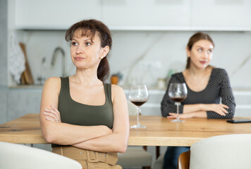 Fototapeta premium Upset middle-aged woman sitting at the kitchen table with her back to another lady quarreling angrily to her