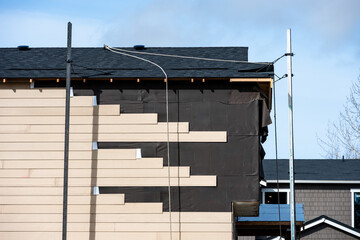New housing construction site, now house with moisture prevention membrane and wood siding being installed with scaffolding, sunny winter day on job site

