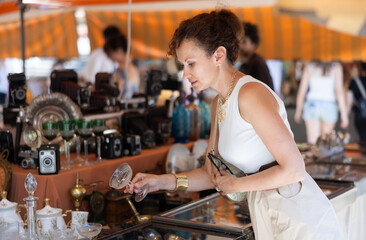 Elegant woman appreciating antiques, browsing with interest vintage glassware at local flea market, searching for unusual item to decorate home or gift..