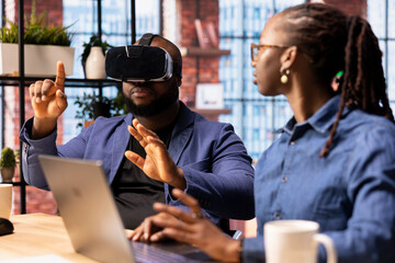 African American man interacts with VR while his girlfriend manages the 3D animation on a laptop at desk. Young couple integrates artificial intelligence and creativity in their workspace.
